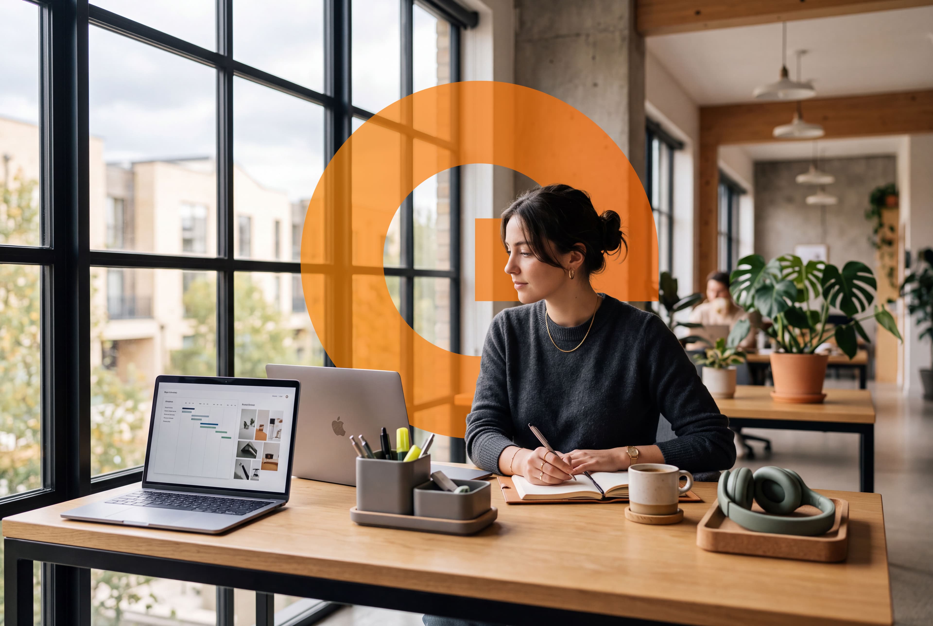 A woman taking notes in her notebook at a wooden desk by the window; two laptops, a pen holder, a coffee cup and headphones on the desk, with the orange esoltec emblem behind her